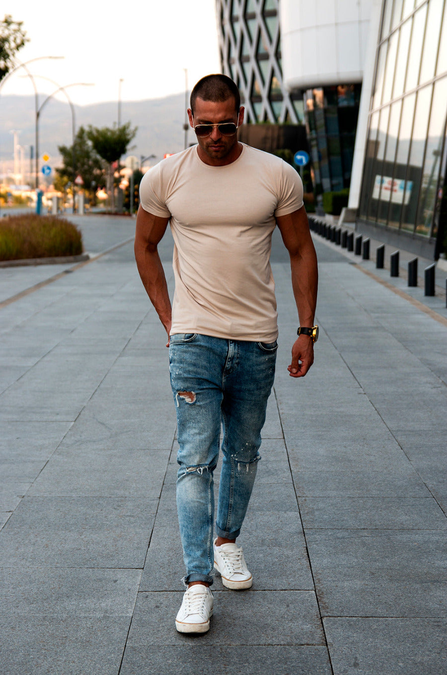 A man wearing a plain light beige crew neck t-shirt and blue jeans, sunglasses, and white shoes, walking on a pavement.